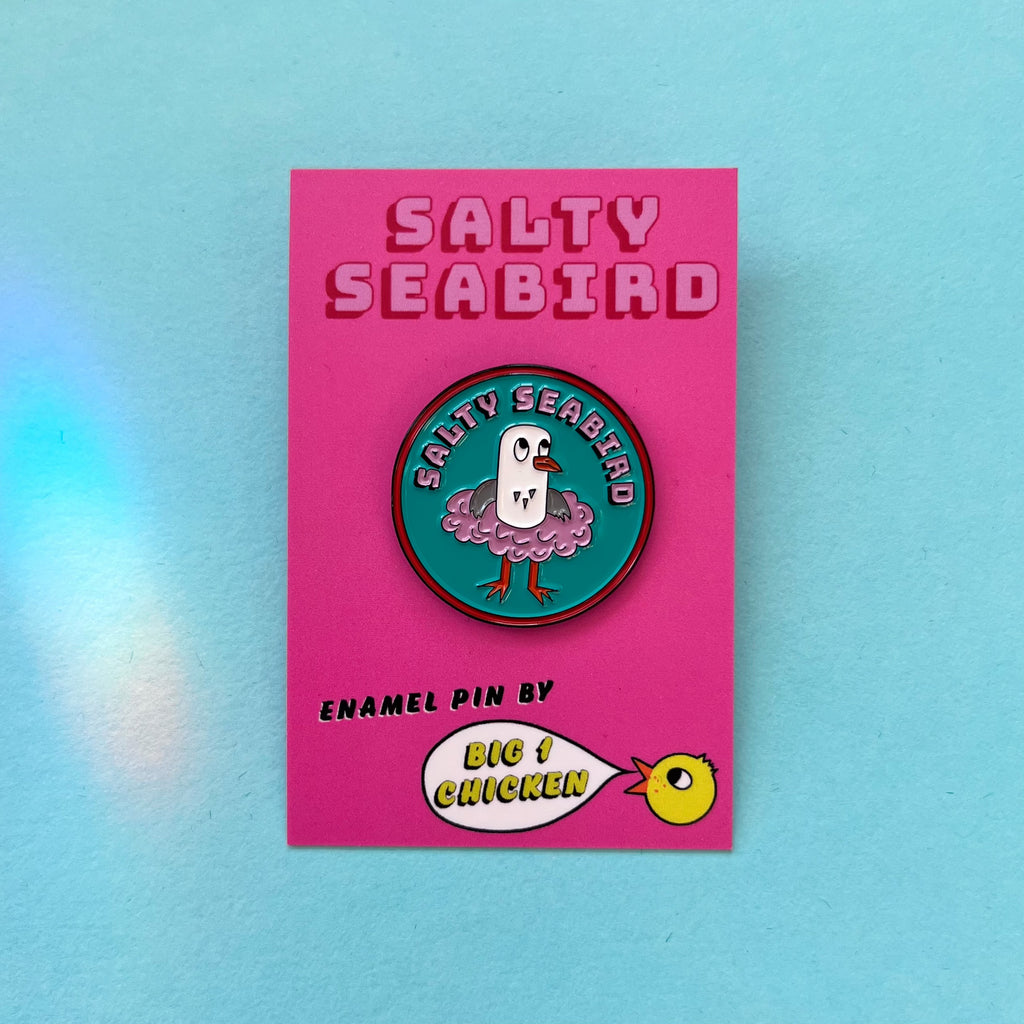 Salty Seabird is a seagull. Here she is seen in her pink tutu on a round enamel pin badge which is approximately 3cm tall. She is on a nice turquoise background. She is grey and white and has an orange beak and orange feet. The pin has pink text on it that says Salty Seabird on pink with a black drop shadow, it has a red line around the circle. The rubber fastener on the back of the pin is a lovely pink colour.She comes on a big 1 chicken card backing with her name on it.