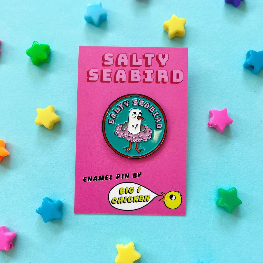 Salty Seabird is a seagull. Here she is seen in her pink tutu on a round enamel pin badge which is approximately 3cm tall. She is on a nice turquoise background. She is grey and white and has an orange beak and orange feet. The pin has pink text on it that says Salty Seabird on pink with a black drop shadow, it has a red line around the circle. The rubber fastener on the back of the pin is a lovely pink colour.She comes on a big 1 chicken card backing with her name on it.