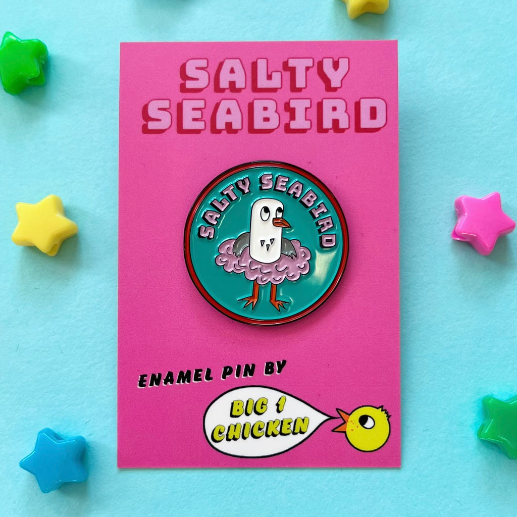 Salty Seabird is a seagull. Here she is seen in her pink tutu on a round enamel pin badge which is approximately 3cm tall.  She is on a nice turquoise background. She is grey and white and has an orange beak and orange feet. The pin has pink text on it that says Salty Seabird on pink with a black drop shadow, it has a red line around the circle. The rubber fastener on the back of the pin is a lovely pink colour. She comes on a big 1 chicken card backing with her name on it.