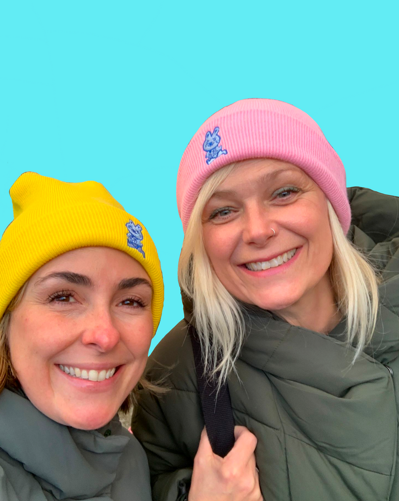 Two happy, smiling women with cute light blue bunny rabbit running with a cheeky look embroidered on their woolly hats.  One cuffed beanie hat is bright yellow and the other is bright pink.