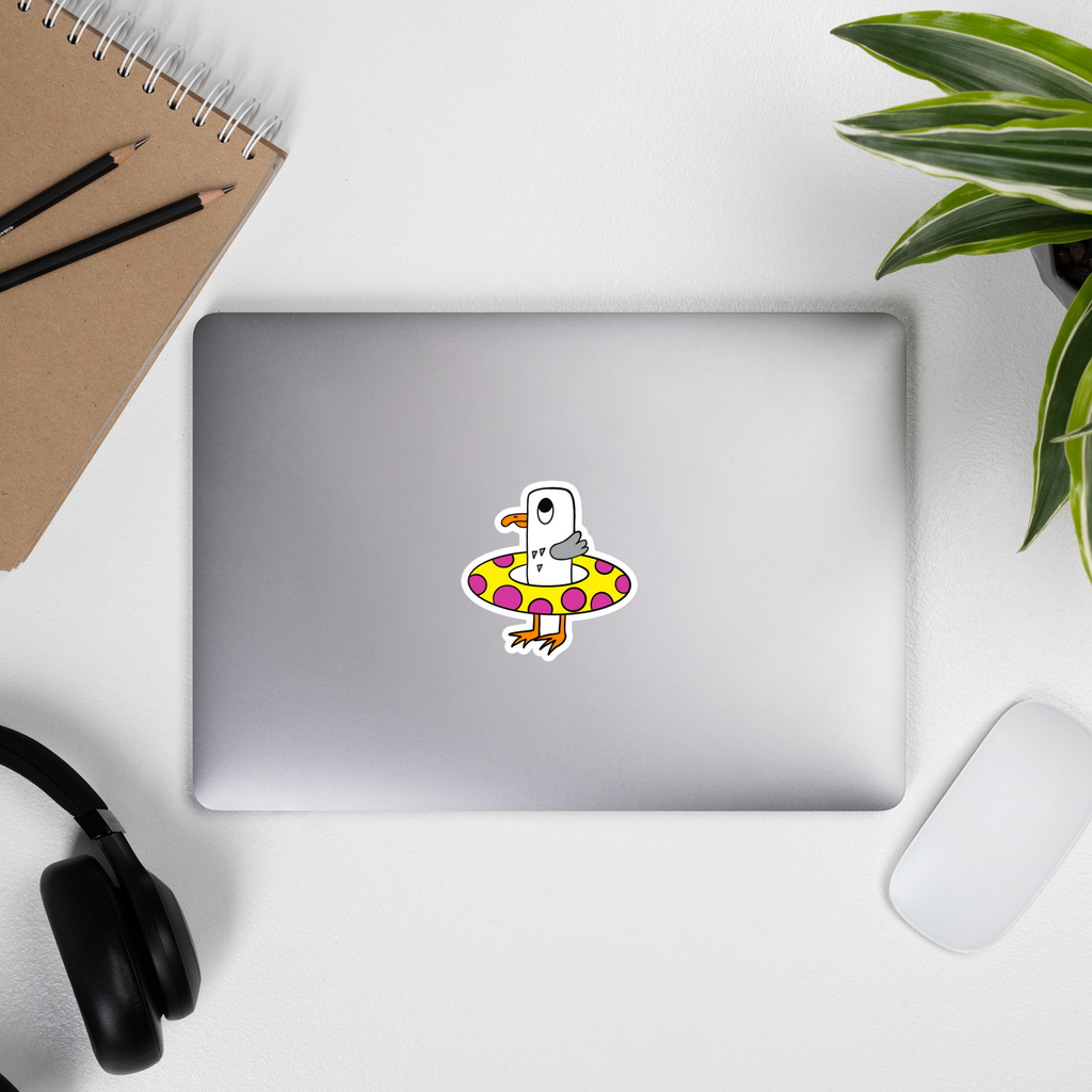 Seagull Steve vinyl, water resistant sticker. Here he can be seen with his bright yellow rubber ring with bright pink circles. Seagull Steve is white and grey with an orange beak and orange feet. He has big wide eyes. Here in this picture you can see Seagull Steve on a silver laptop.